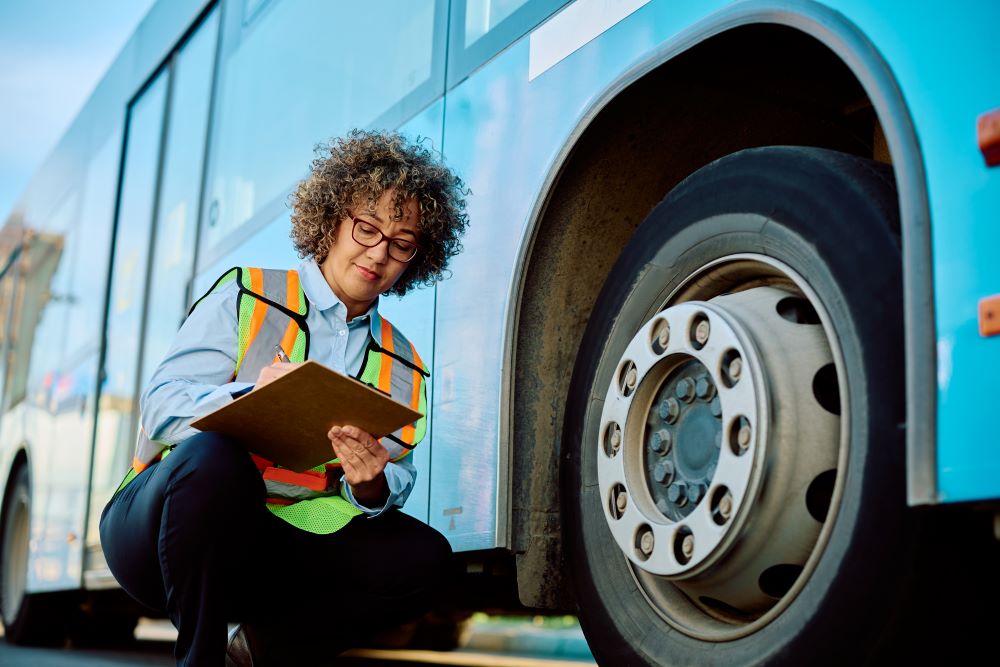 An image showing a woman wearing a safety vest kneeling next to a bus tire. She is inspecting a tire before departure, reflecting proper bus operational standards that preserve the safety of passengers.