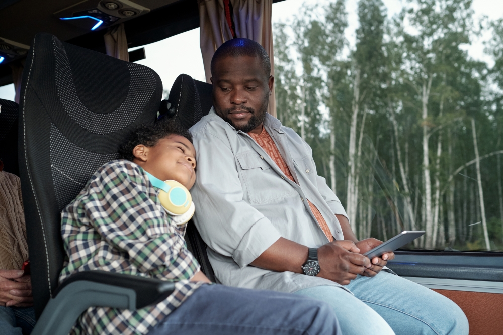 A father and child are seen traveling by bus. The child, wearing a plaid shirt and headphones, sleeps against the bus seat. The adult, wearing a light grey shirt and jeans, looks down at a tablet. Outside the bus windows, birch trees stand in view.