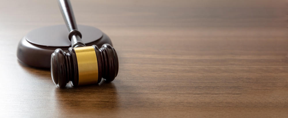 A shiny wooden gavel sits on a dark, textured wooden table in this eye-level shot. The gavel is dark brown with a golden band and resting on a circular base. The table is light brown and wood grain can be seen.