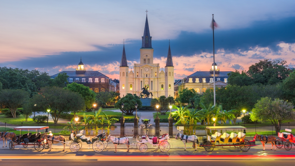 A wide shot depicts a tranquil cityscape featuring a prominent cathedral with spires set against a beautiful twilight sky. Horse-drawn carriages line the street in front of the cathedral and a park with lush green trees separates the street from the building. The photograph captures the transition between day and night, with light trails from vehicles adding a dynamic element.