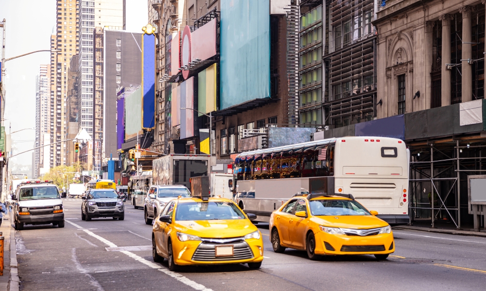 Cars and buses traverse a busy urban street lined with tall buildings and colorful billboards. A single figure is visible walking along the sidewalk, adding a human element to the scene. The daytime lighting highlights the vibrant colors of the city, while the towering buildings and bustling traffic create a sense of energy and movement.